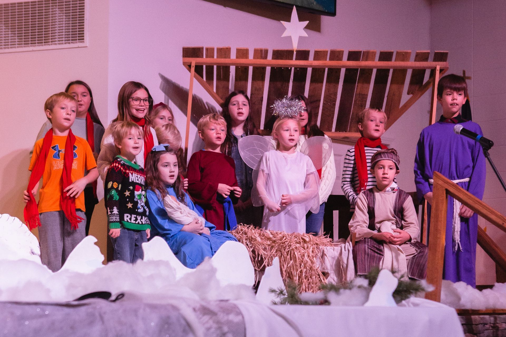 Children performing a Nativity play on stage, singing near a manger set.