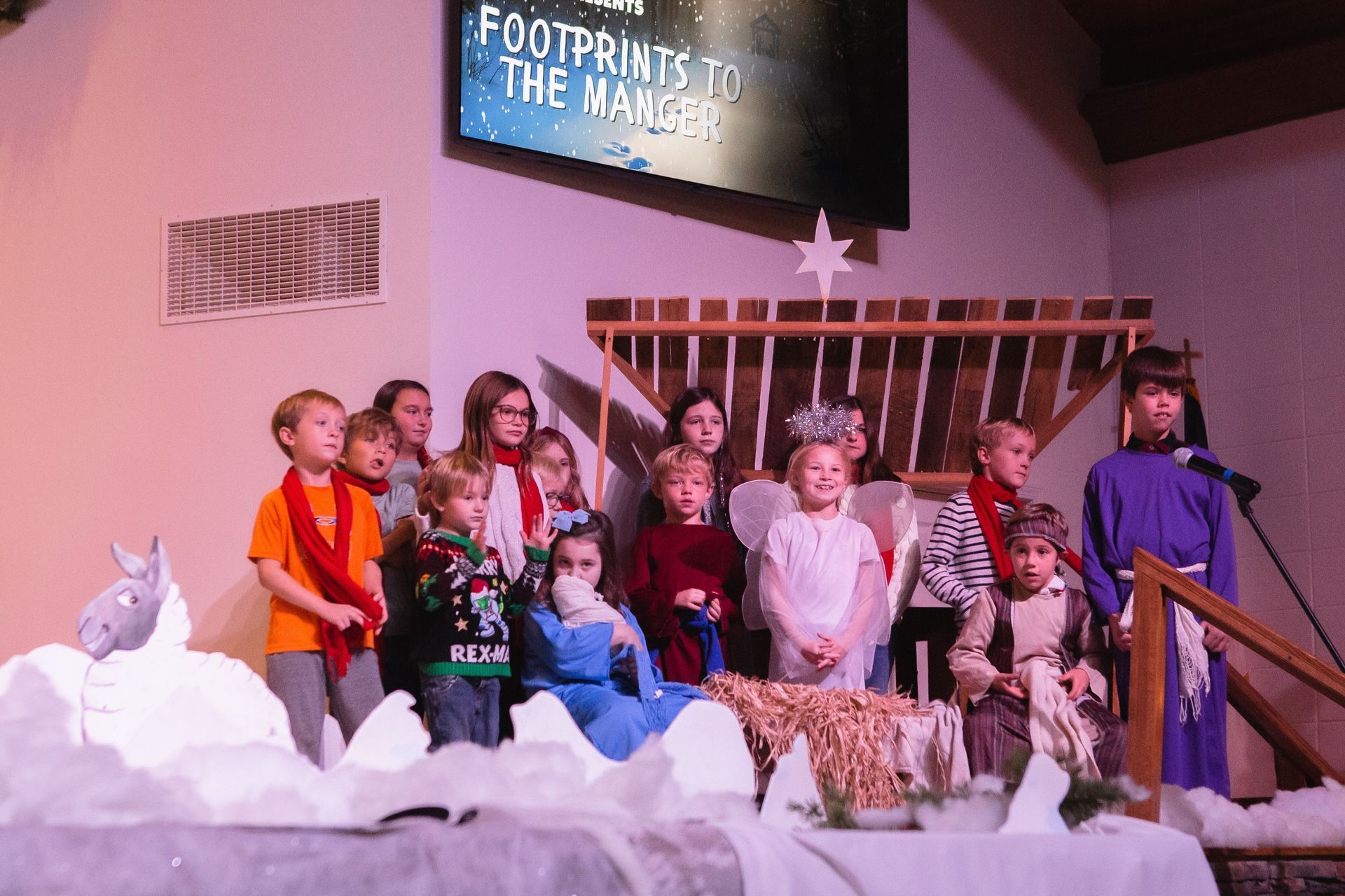 Children performing a nativity play with costumes, props, and a banner reading 
