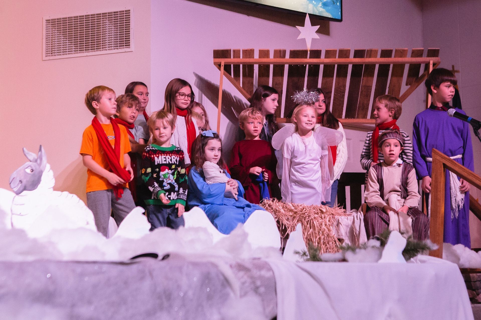 Children performing a Nativity play on a stage, some in costumes, in front of a manger scene.