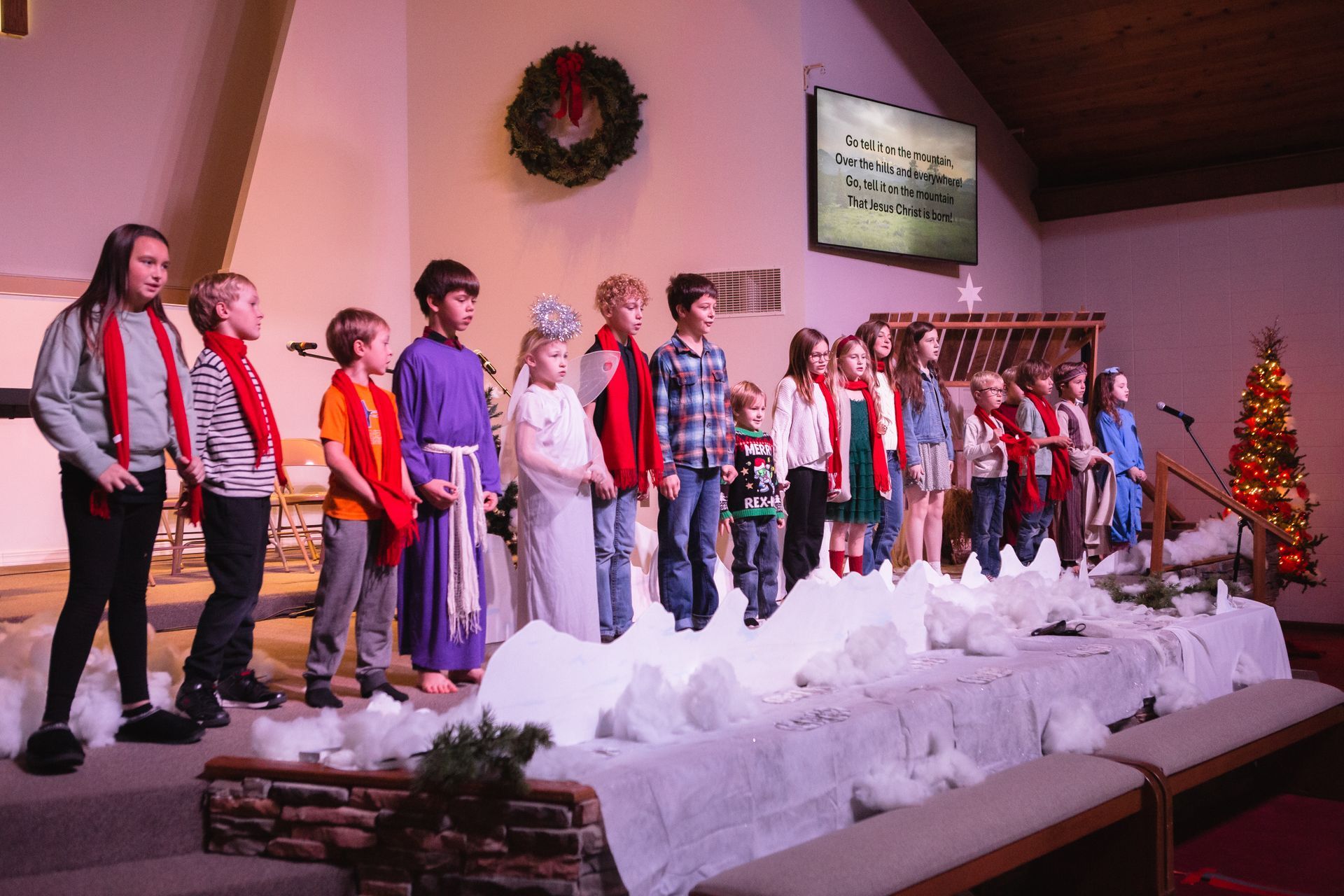 Children's choir on stage, singing. Decorated with snow and Christmas tree.