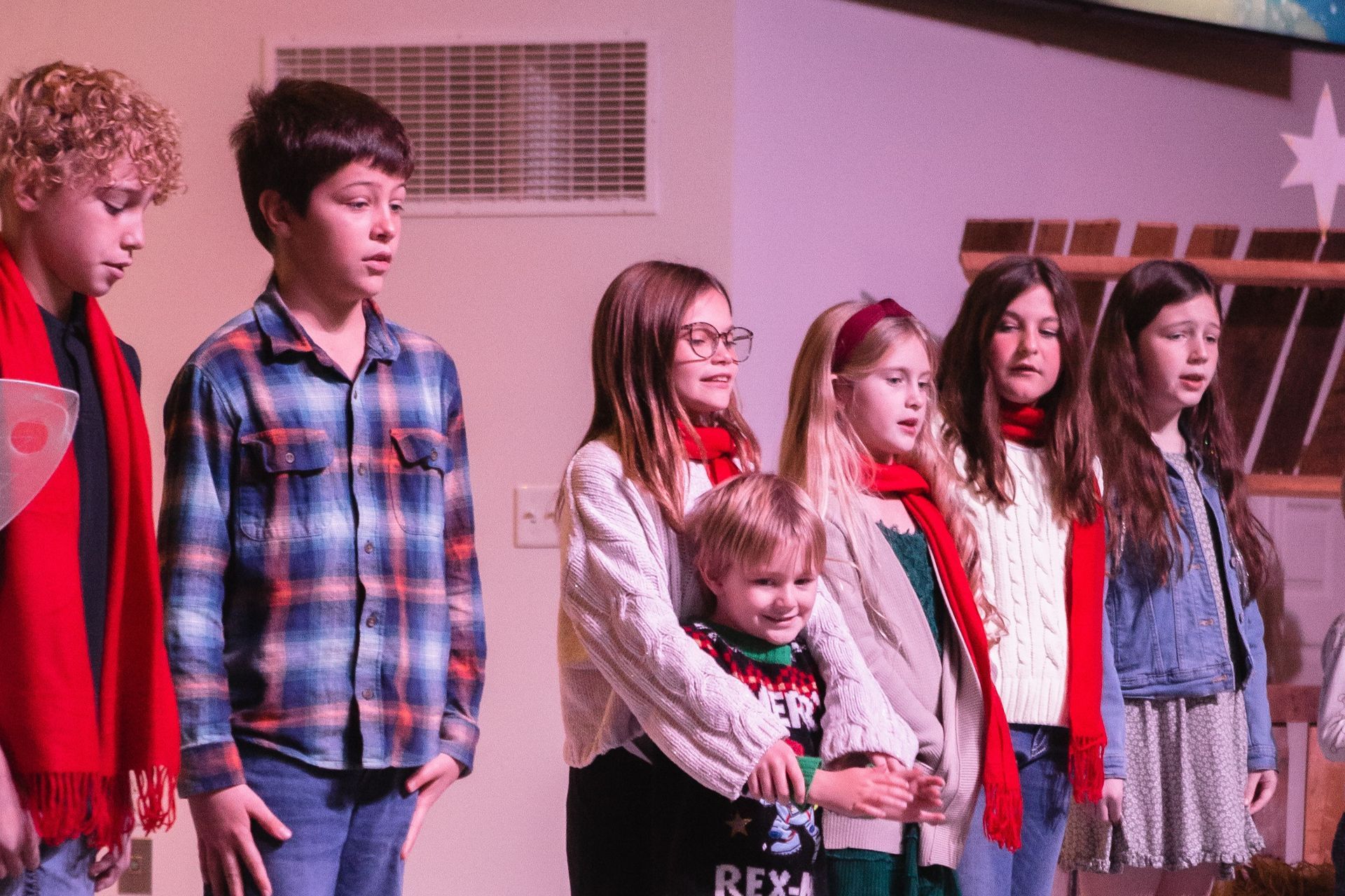 Children singing on stage, wearing scarves and sweaters; some are looking forward.