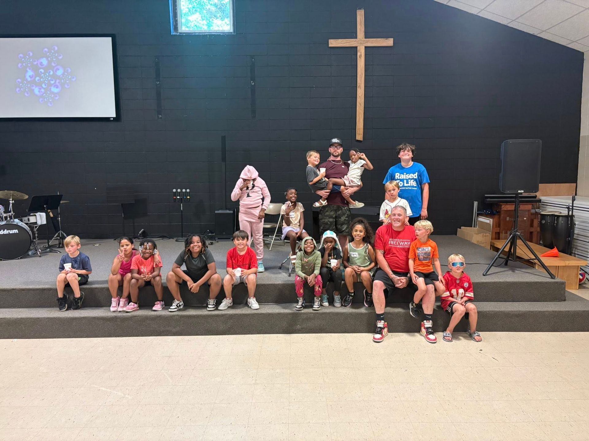 A group of children are sitting on a stage in front of a cross.