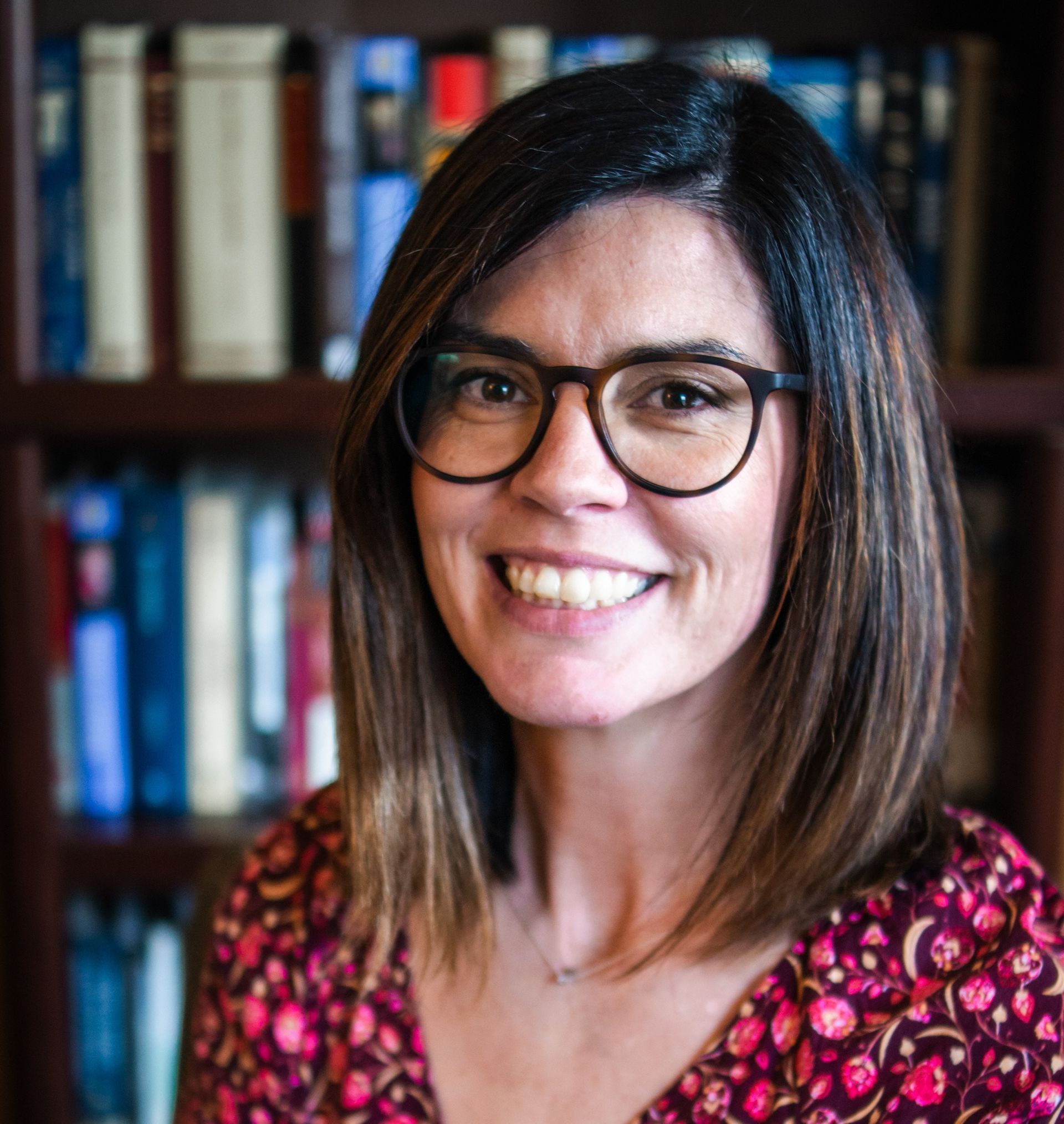 A woman wearing glasses is smiling in front of a bookshelf.