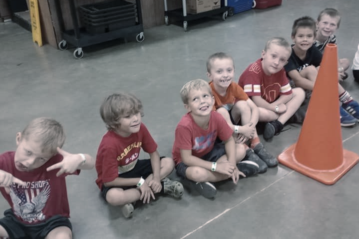 A group of young boys sitting on the floor in a line, with an orange cone nearby.