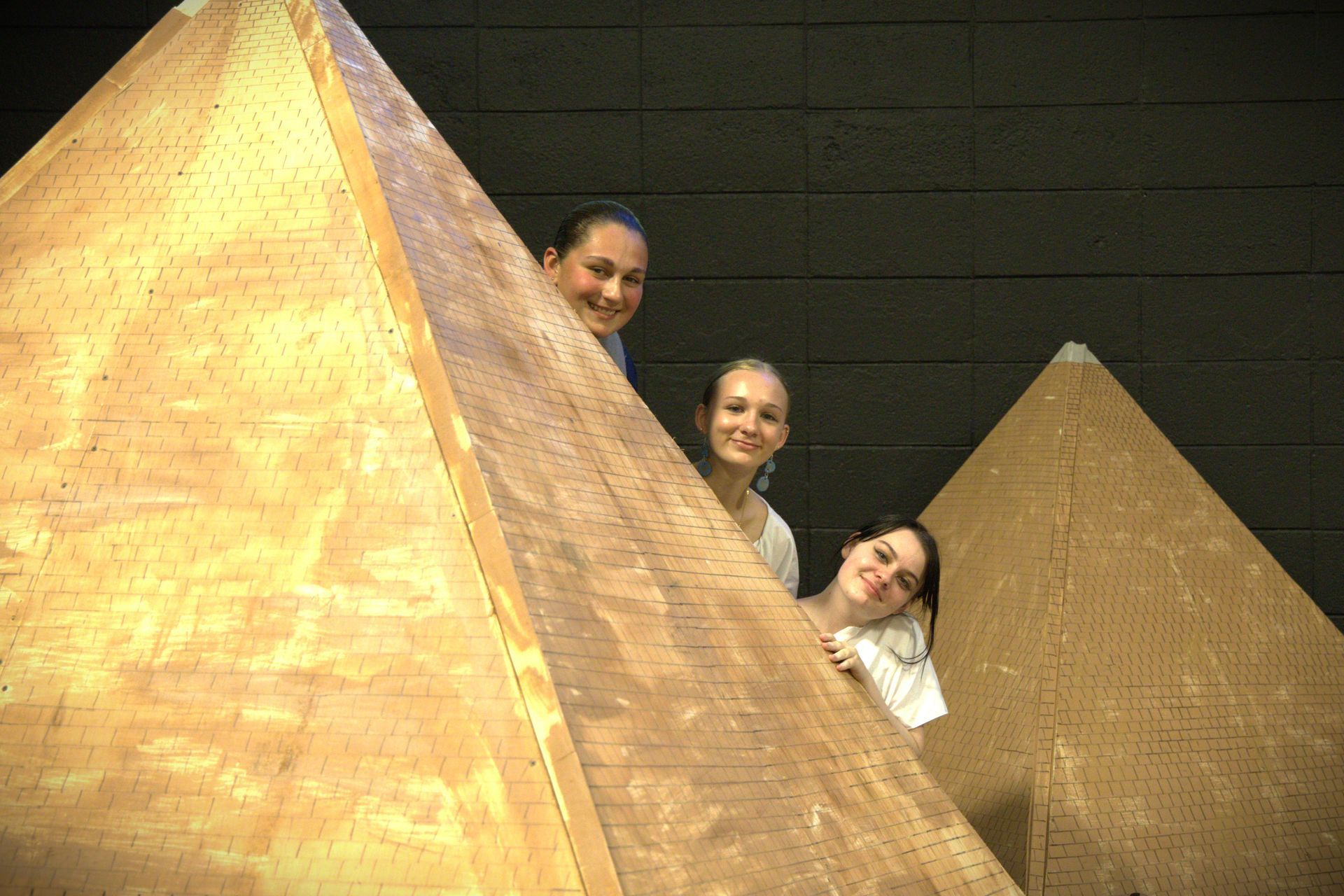 Three women are peeking out from behind a pyramid.