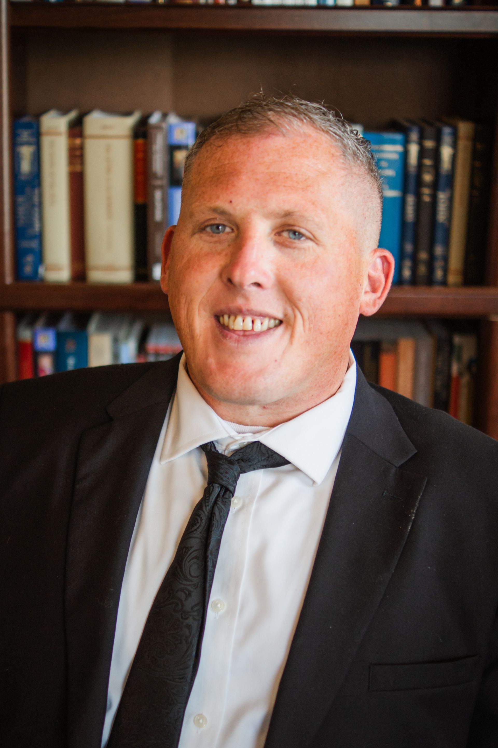A man in a suit and tie is smiling in front of a bookshelf.