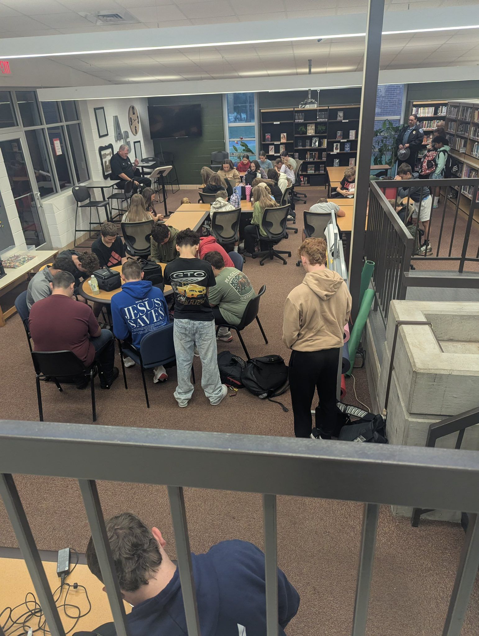 Students gather around tables in a library. Some are seated, others stand. Fluorescent lights illuminate the room.
