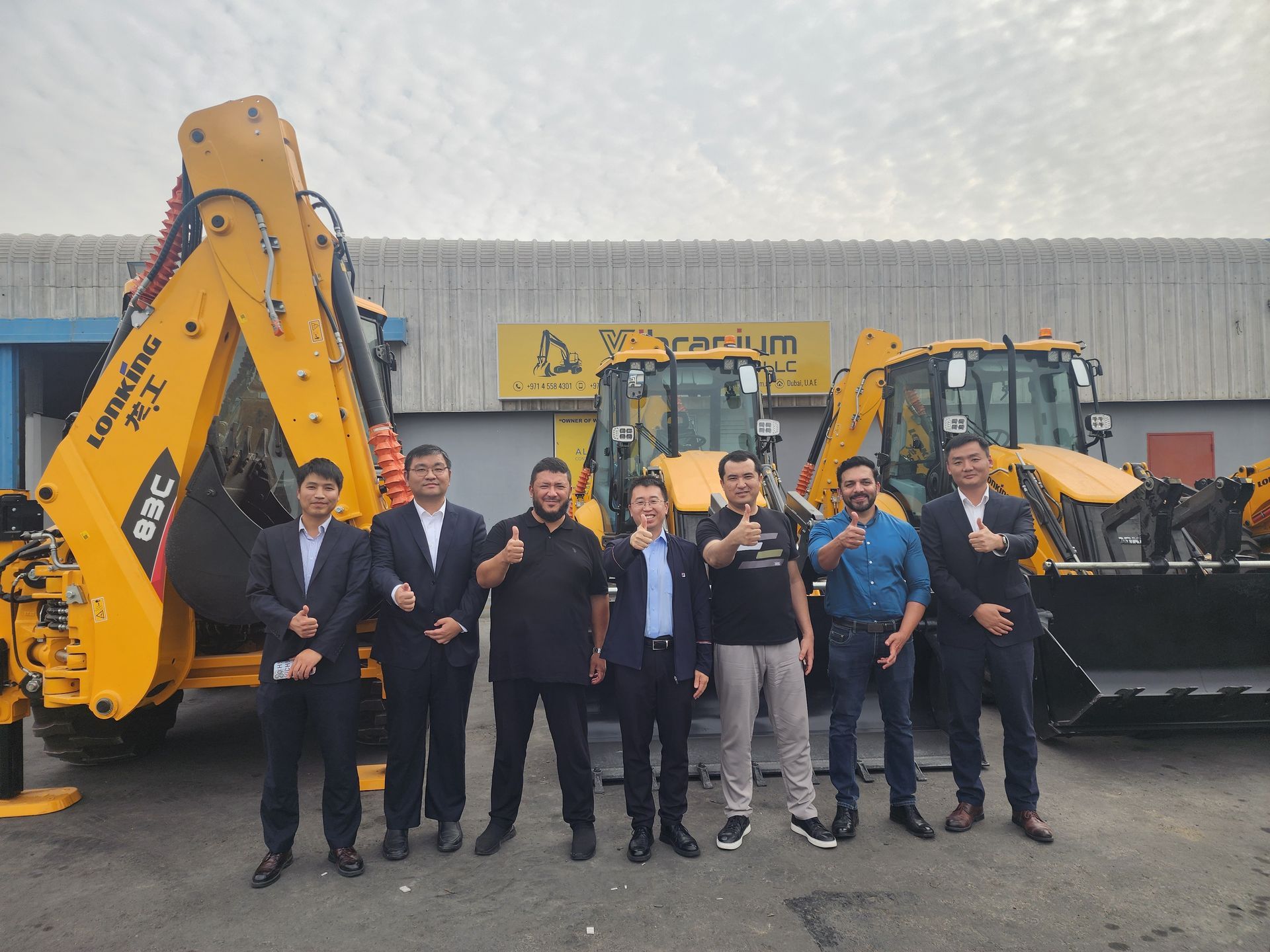 A group of men standing in front of yellow construction vehicles. They are outdoors on a cloudy day, many with thumbs up.