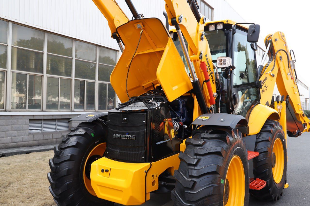 Yellow backhoe with raised engine compartment, parked outside a building.