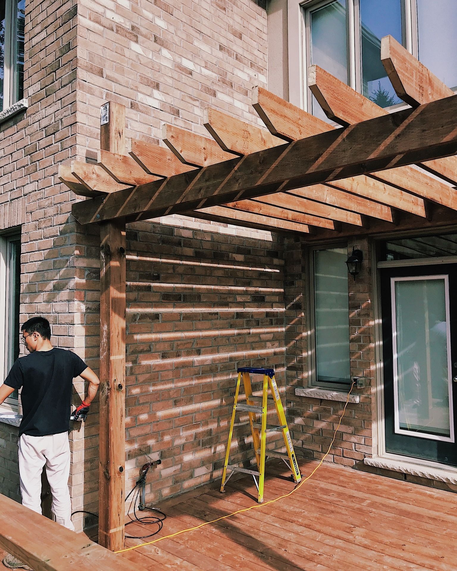 A man is working on a wooden pergola on the side of a brick building.
