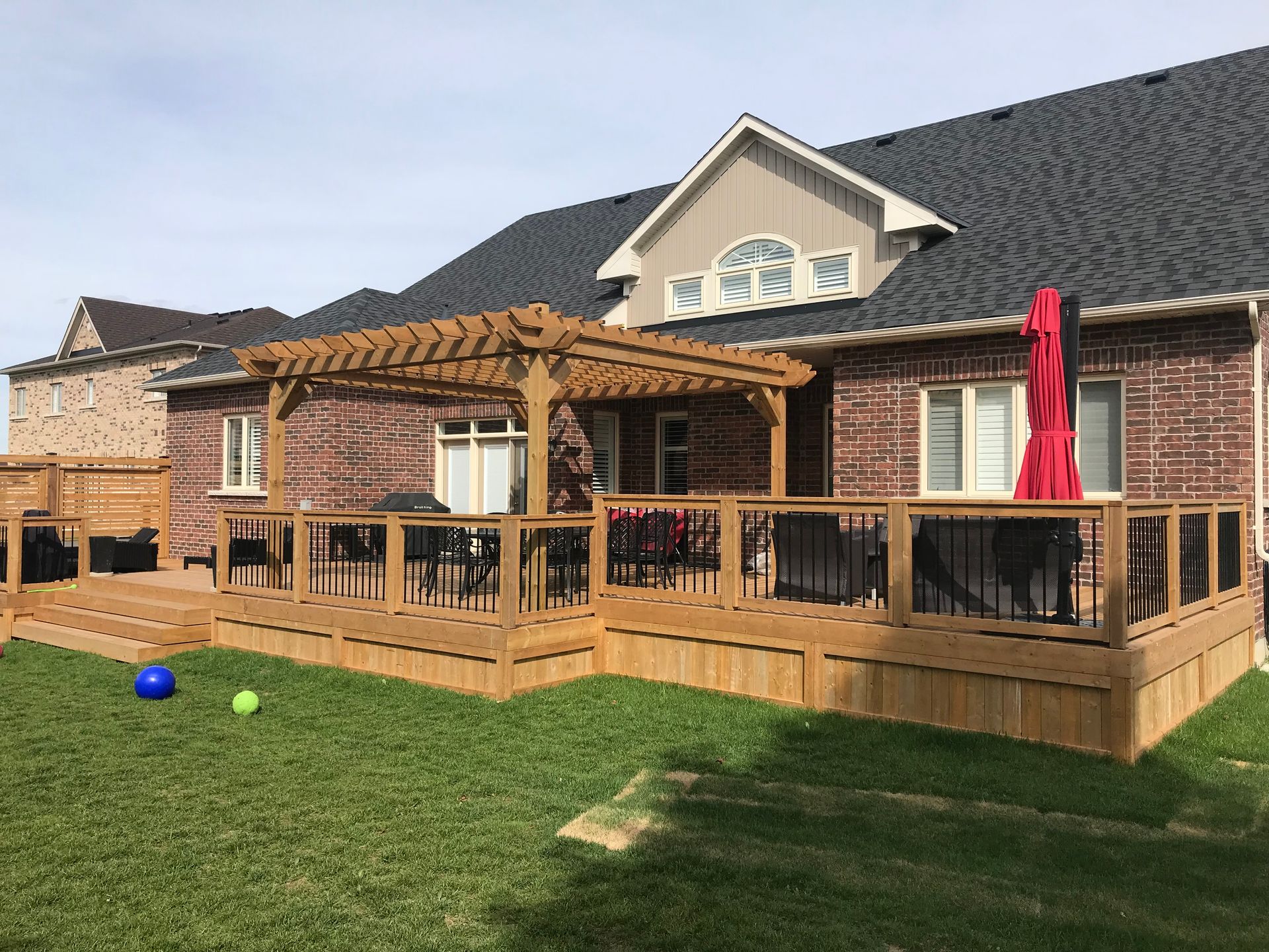 A large wooden deck with a pergola and umbrella in front of a brick house.