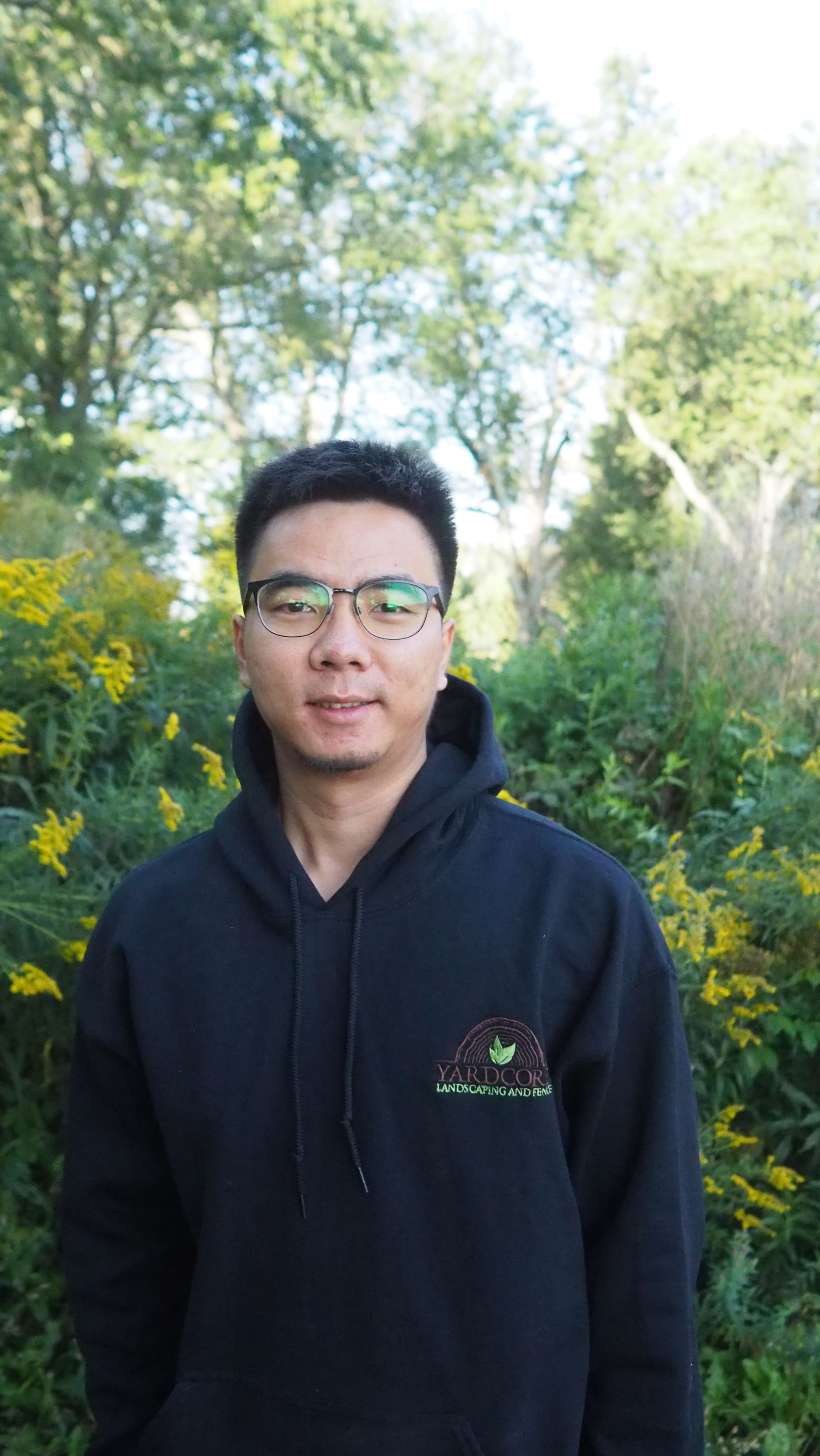 A man wearing glasses and a black hoodie is standing in front of a bush.