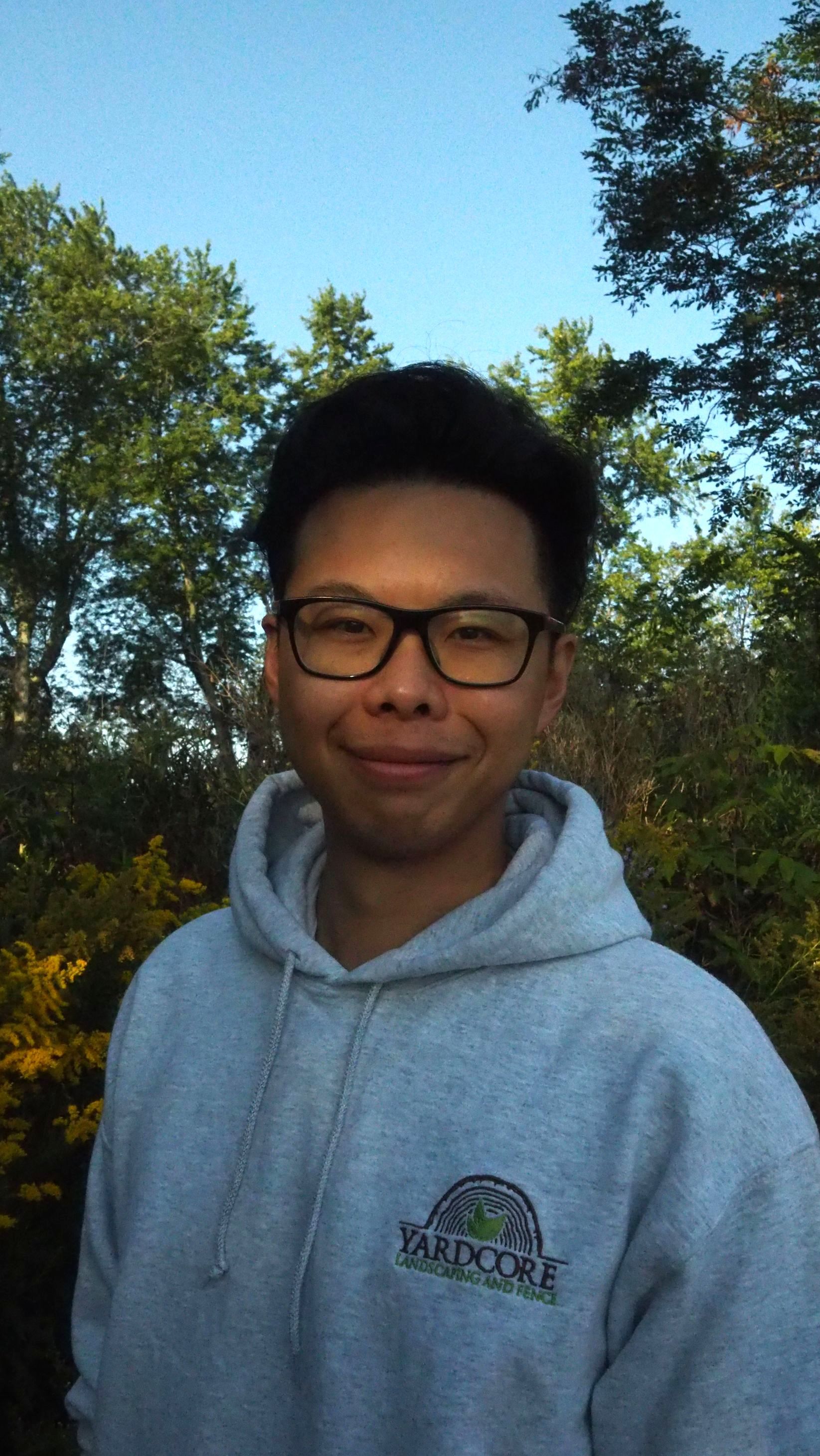 A young man wearing glasses and a hoodie is standing in front of trees.