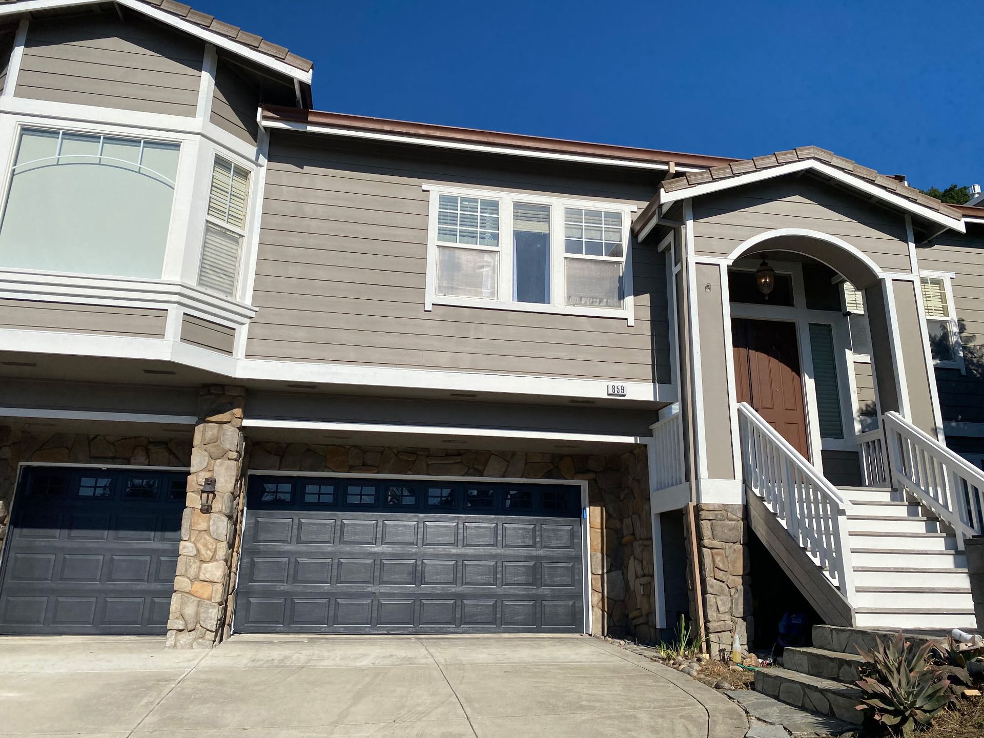 Two-story house with gray siding, stone accents, dark garage doors, and white trim against a blue sky.