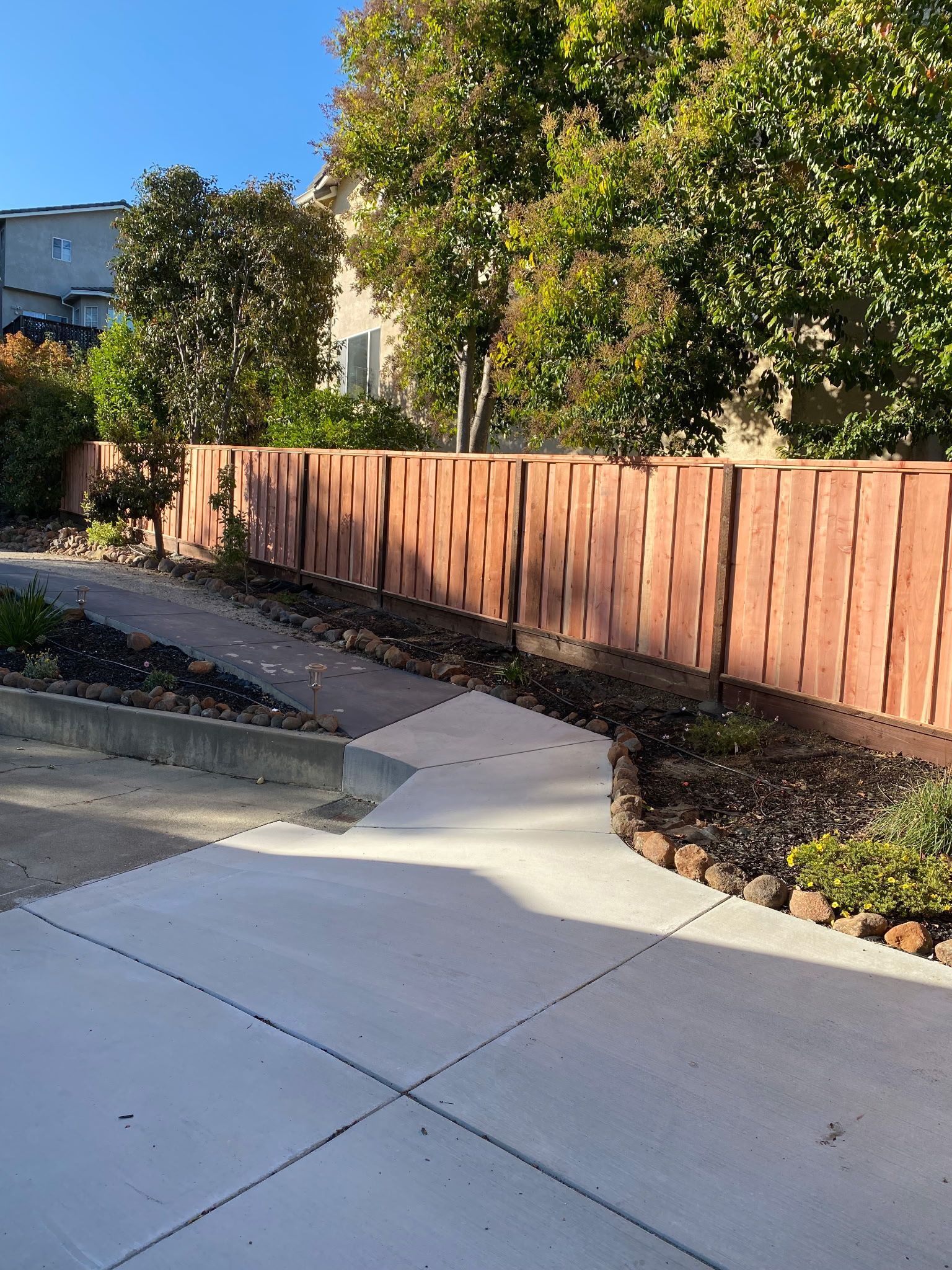 Concrete walkway leads to a textured brown wall, flanked by trees and greenery.
