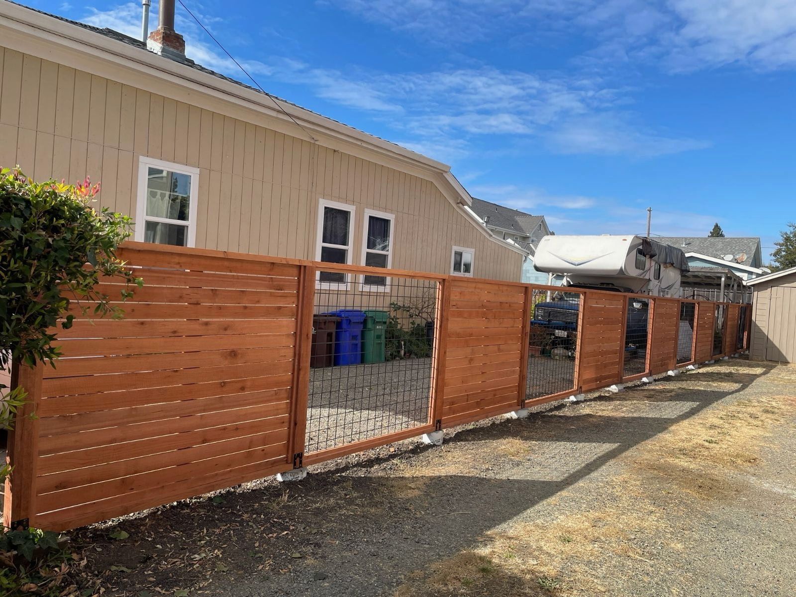 Wooden fence along a building, gravel ground, blue sky, parked car visible.