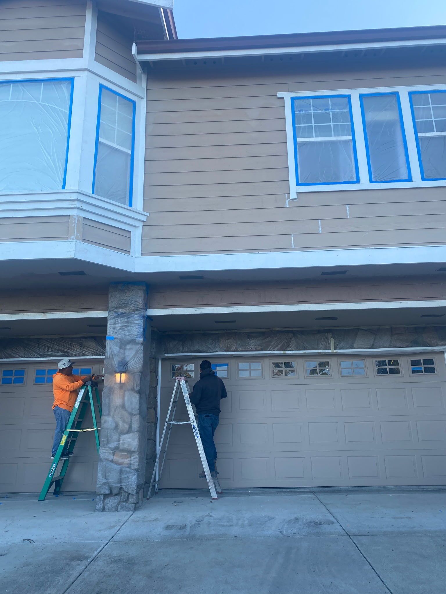 Two people painting a house. Garage doors and windows taped with blue painter's tape. Beige house with brown trim.