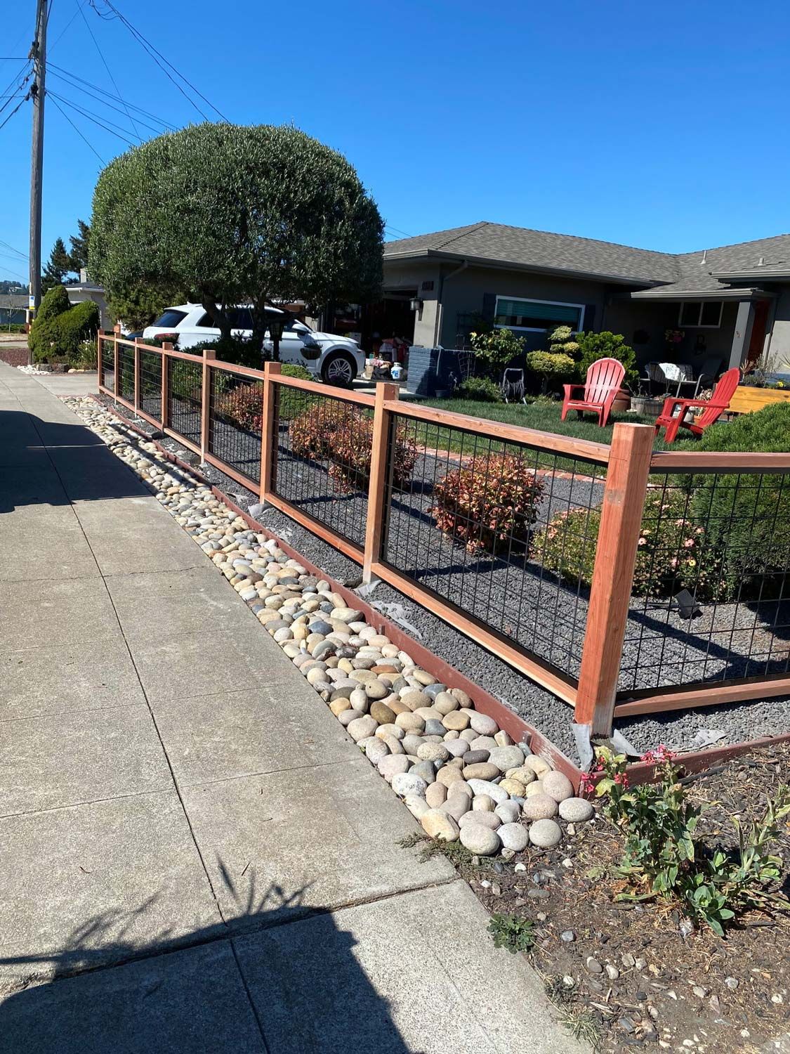 Wooden fence with wire mesh bordering a sidewalk and yard with plants and rocks.