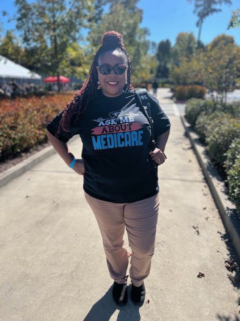 A woman wearing a black t-shirt that says `` medicare '' is standing on a sidewalk.