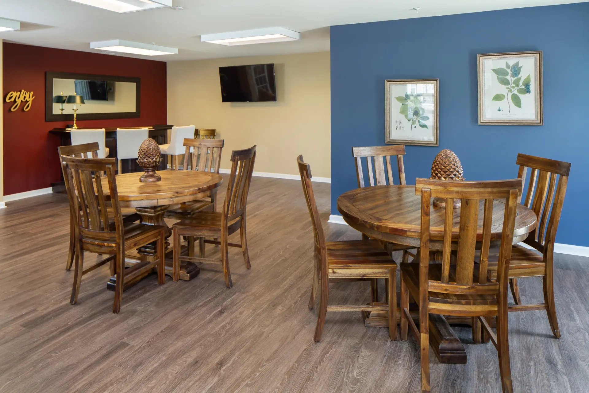 Indoor community lounge with two round wooden tables and wooden chairs, blue accent wall and wall art.