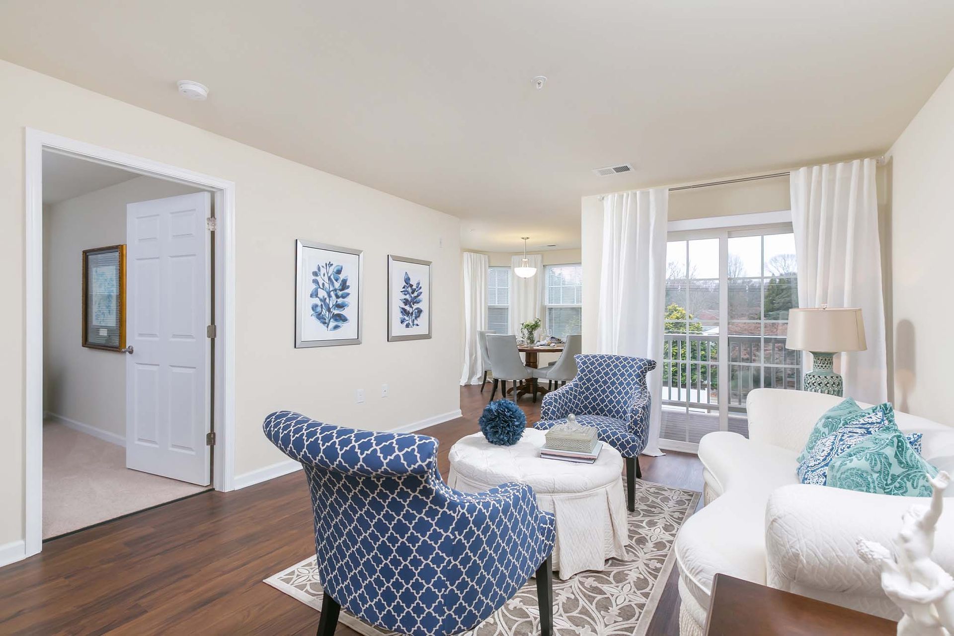 Living room with blue patterned chairs around a round ottoman, white sofa, and a balcony with white curtains.