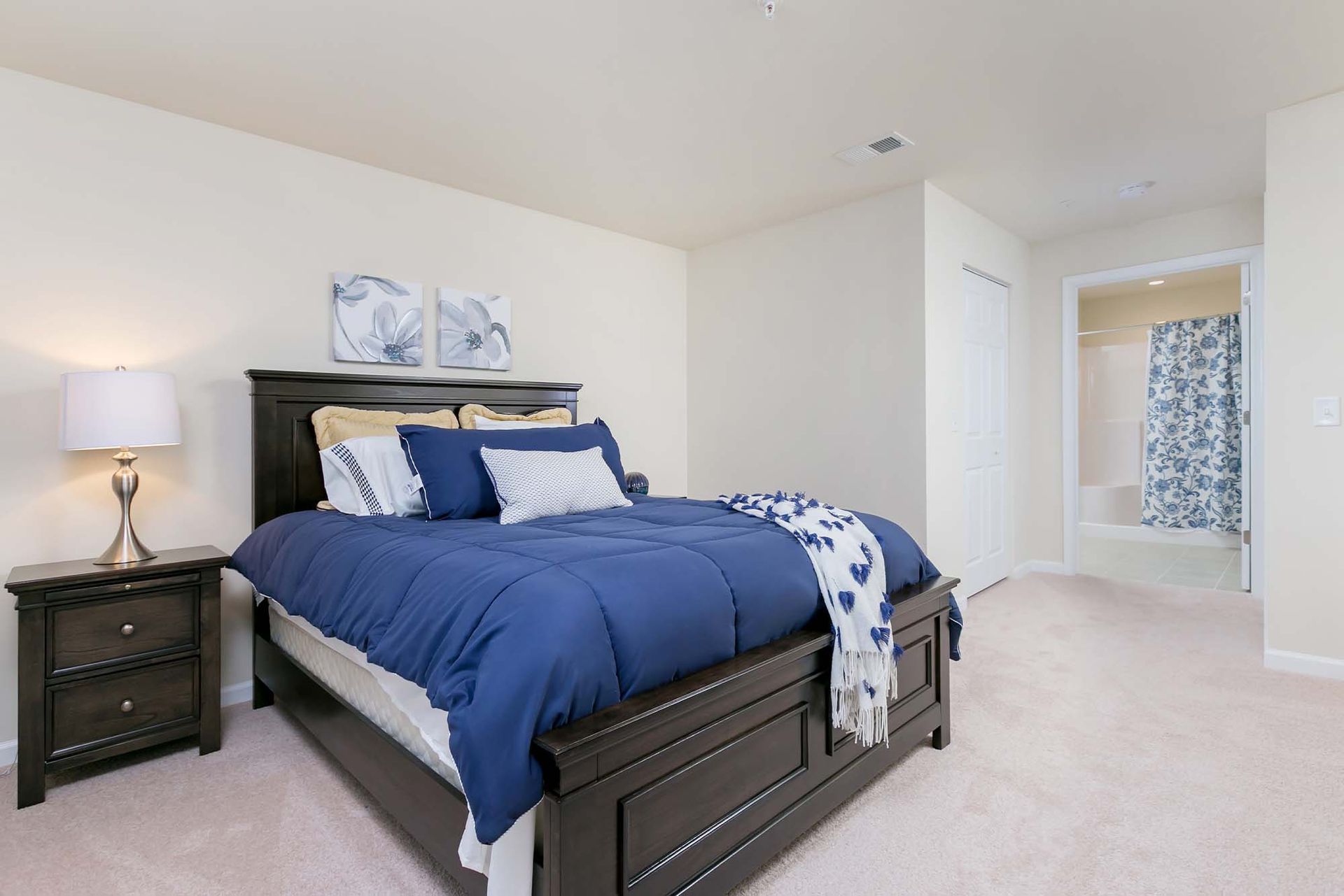 Bedroom with dark wood bed, navy bedding, a nightstand lamp, and an en-suite bathroom with a blue floral shower curtain.