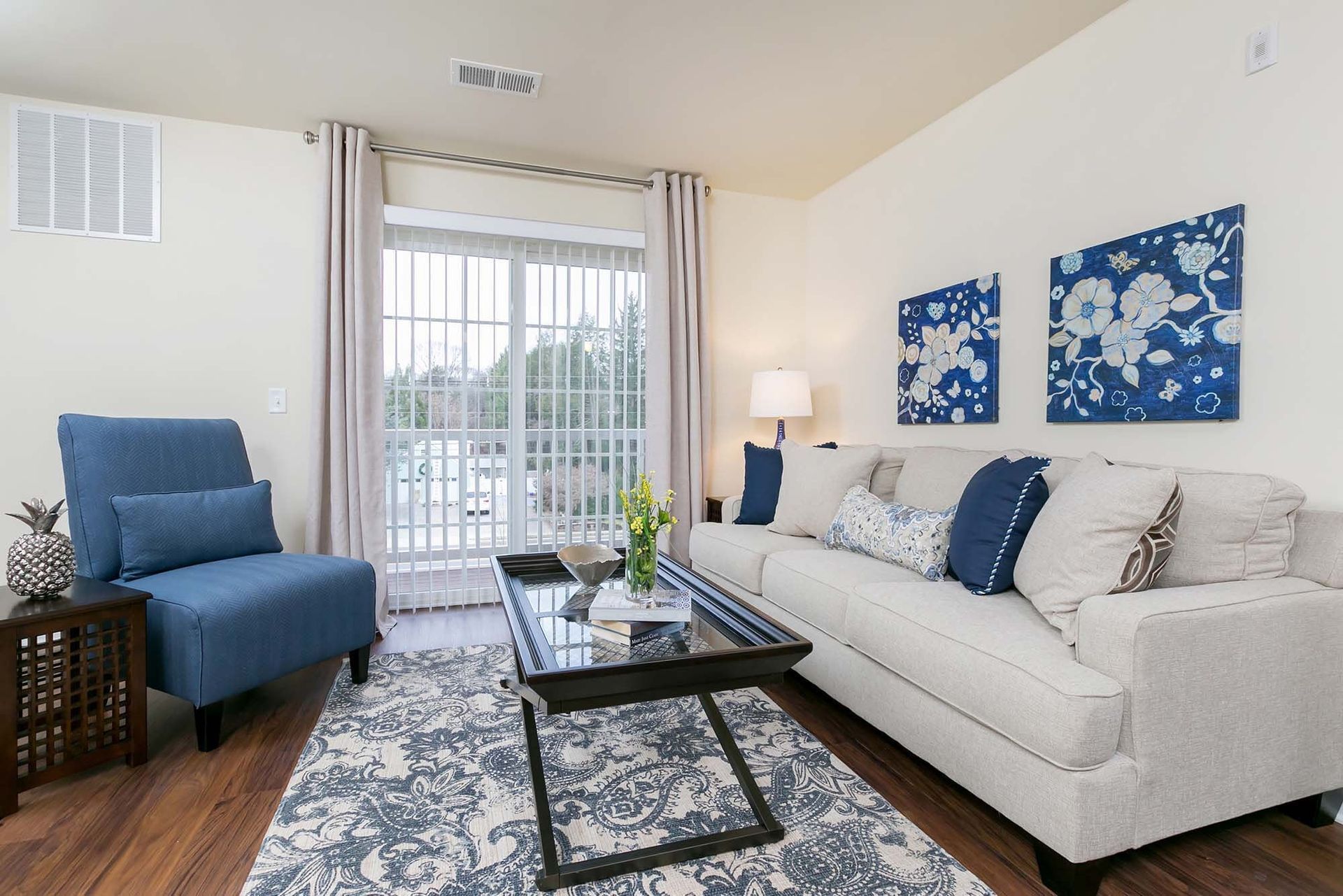 Bright living room with beige walls, blue accent chair, light sofa, coffee table, and sliding glass door to balcony.