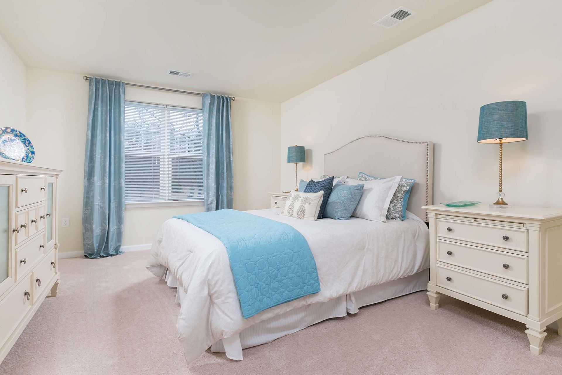 Cozy apartment bedroom with white furniture, a tufted headboard, blue bedding, and matching lamps.