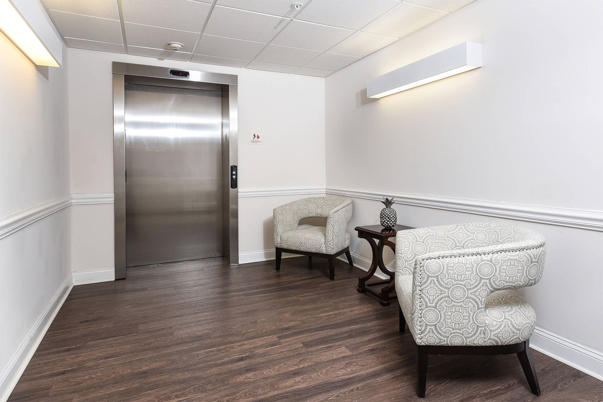 Elevator lobby with a stainless steel elevator, two patterned armchairs, and a small side table.