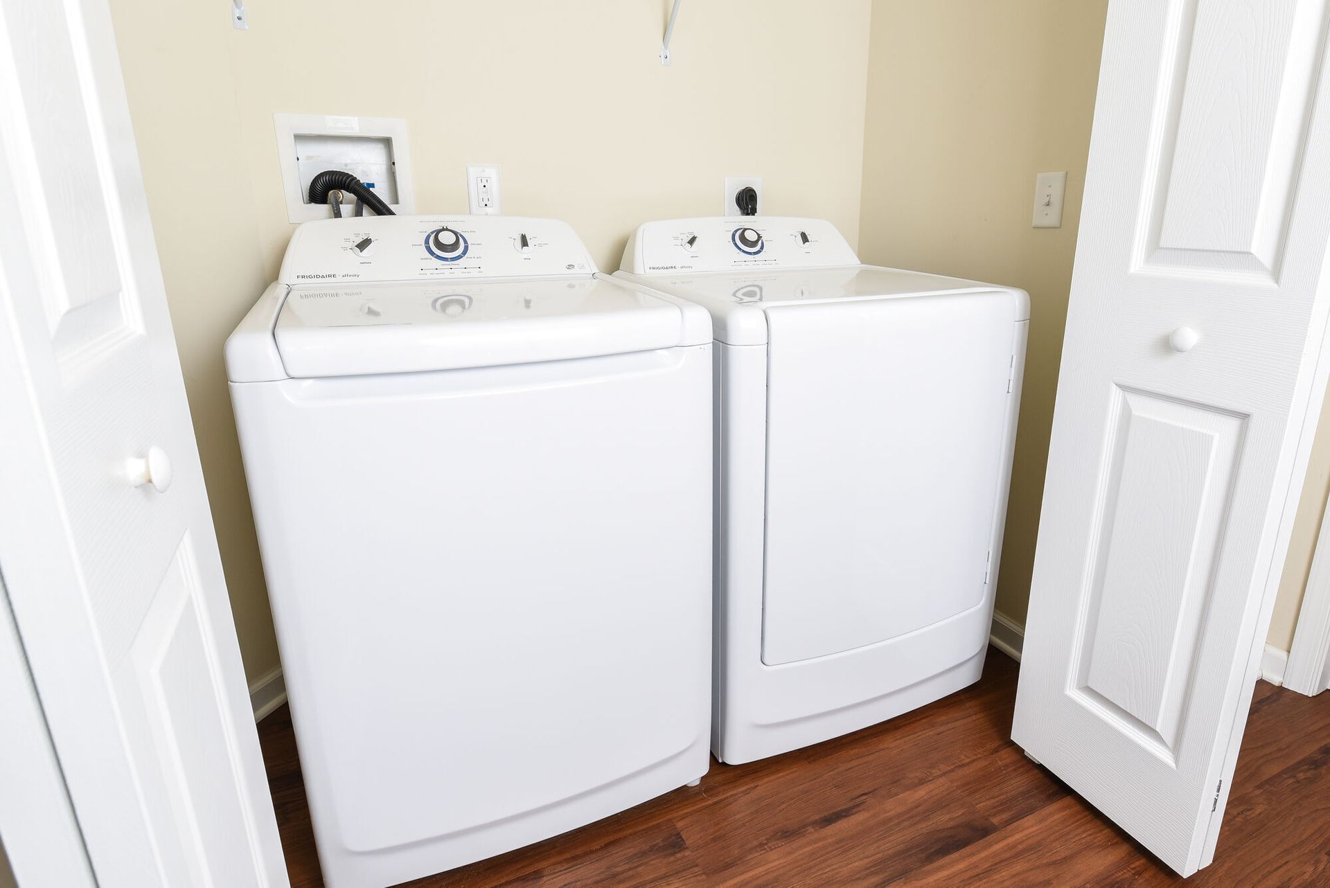 Two white front-loading laundry appliances in a compact beige closet with wood flooring.