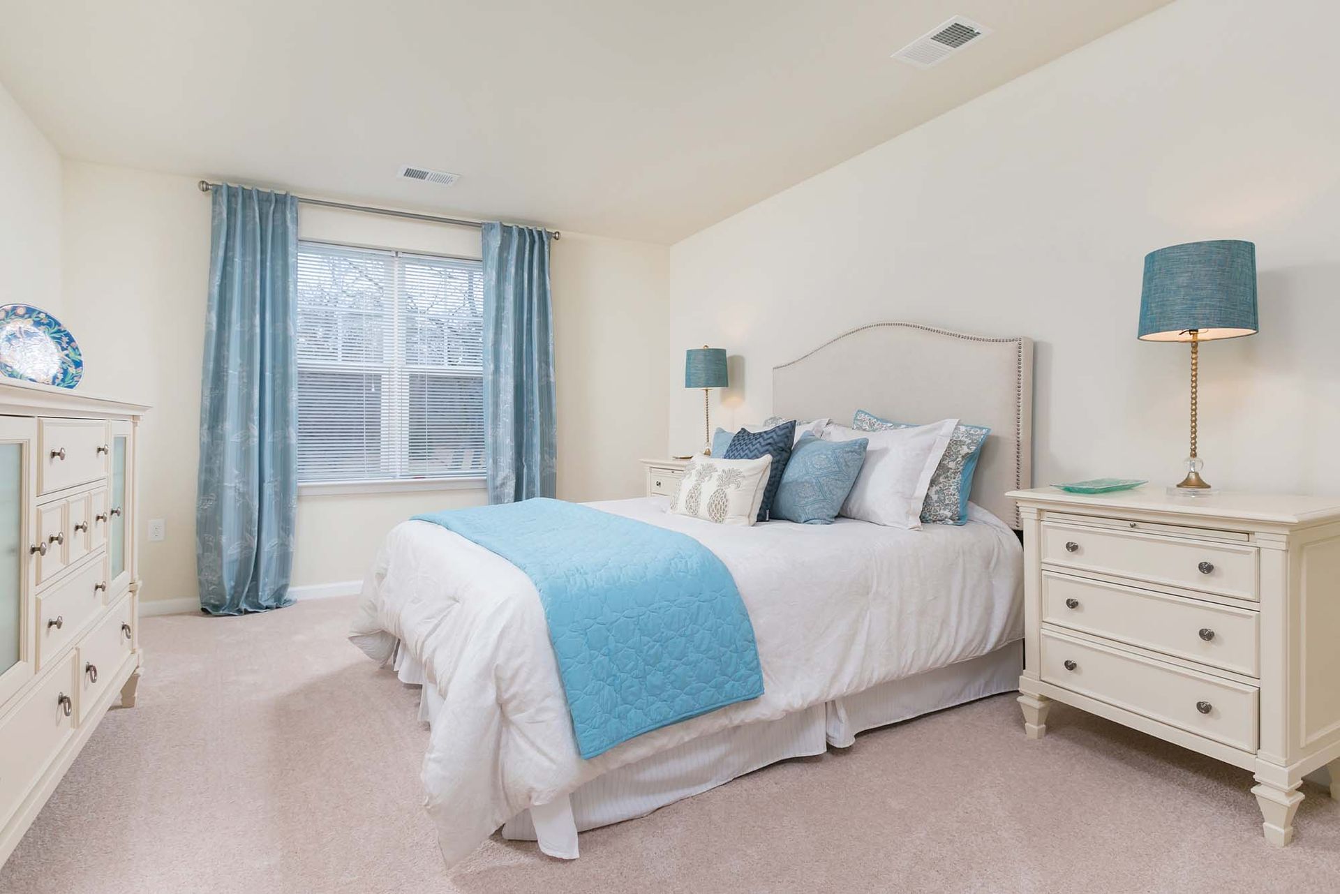 Bedroom in apartment with white furniture, blue bedding, and a large window with blue curtains.
