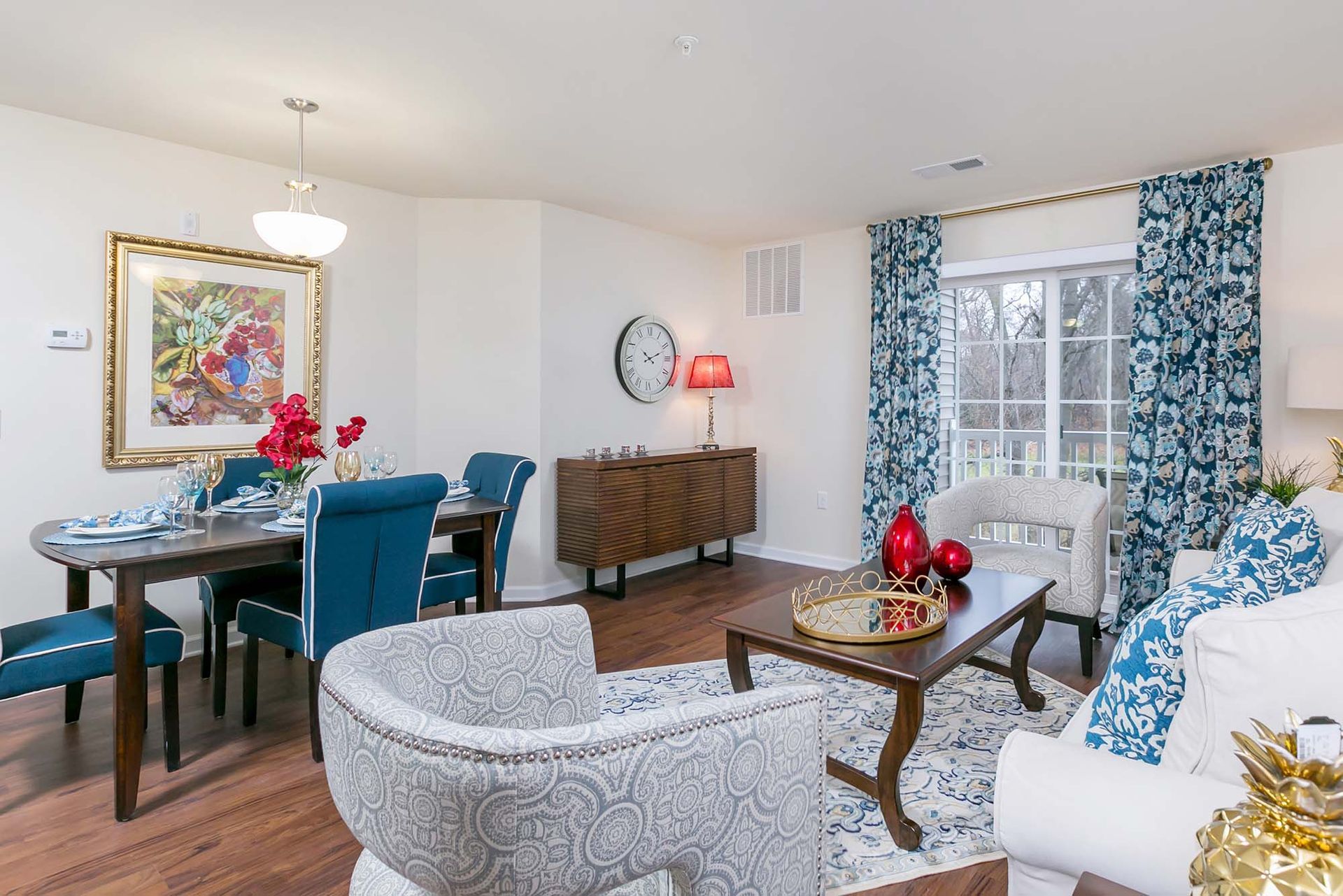 Living and dining area of an apartment with blue upholstered chairs, floral curtains, and a balcony door.