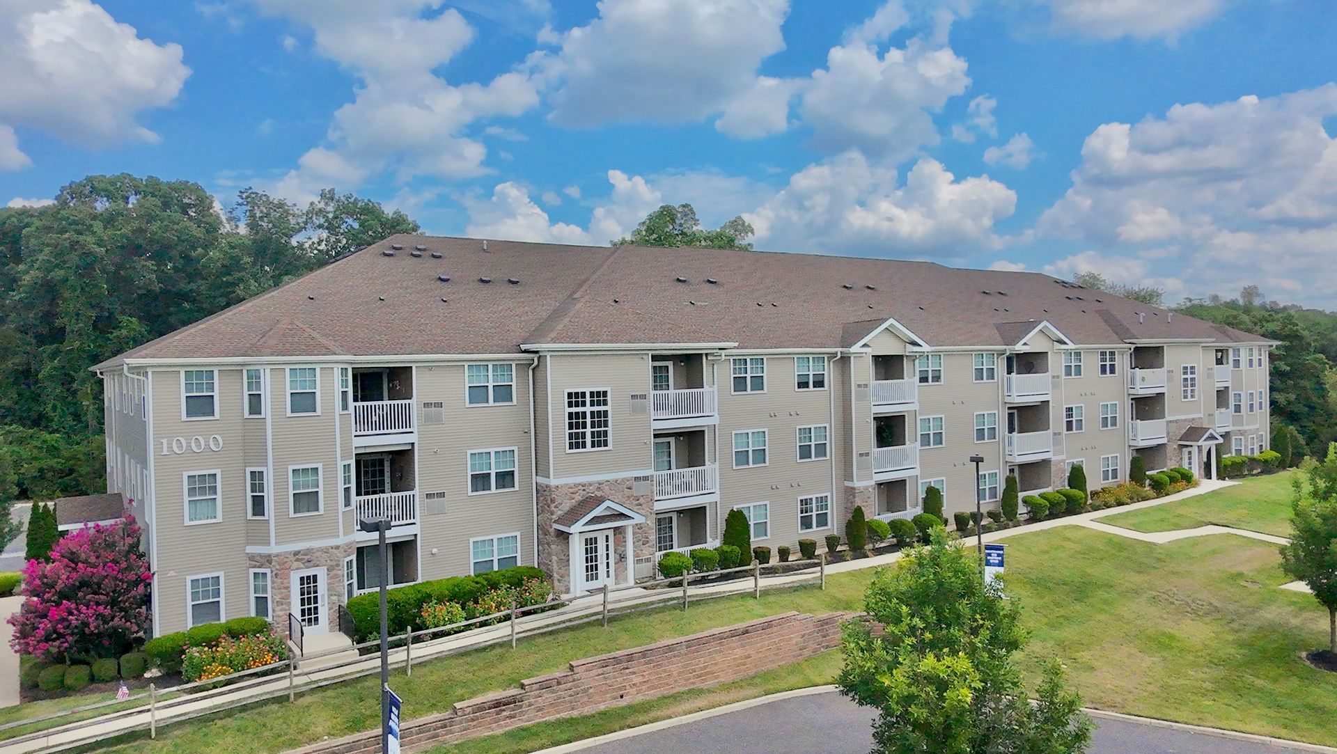 Apartment building with beige siding, brown roof, and balconies under a blue sky with clouds.