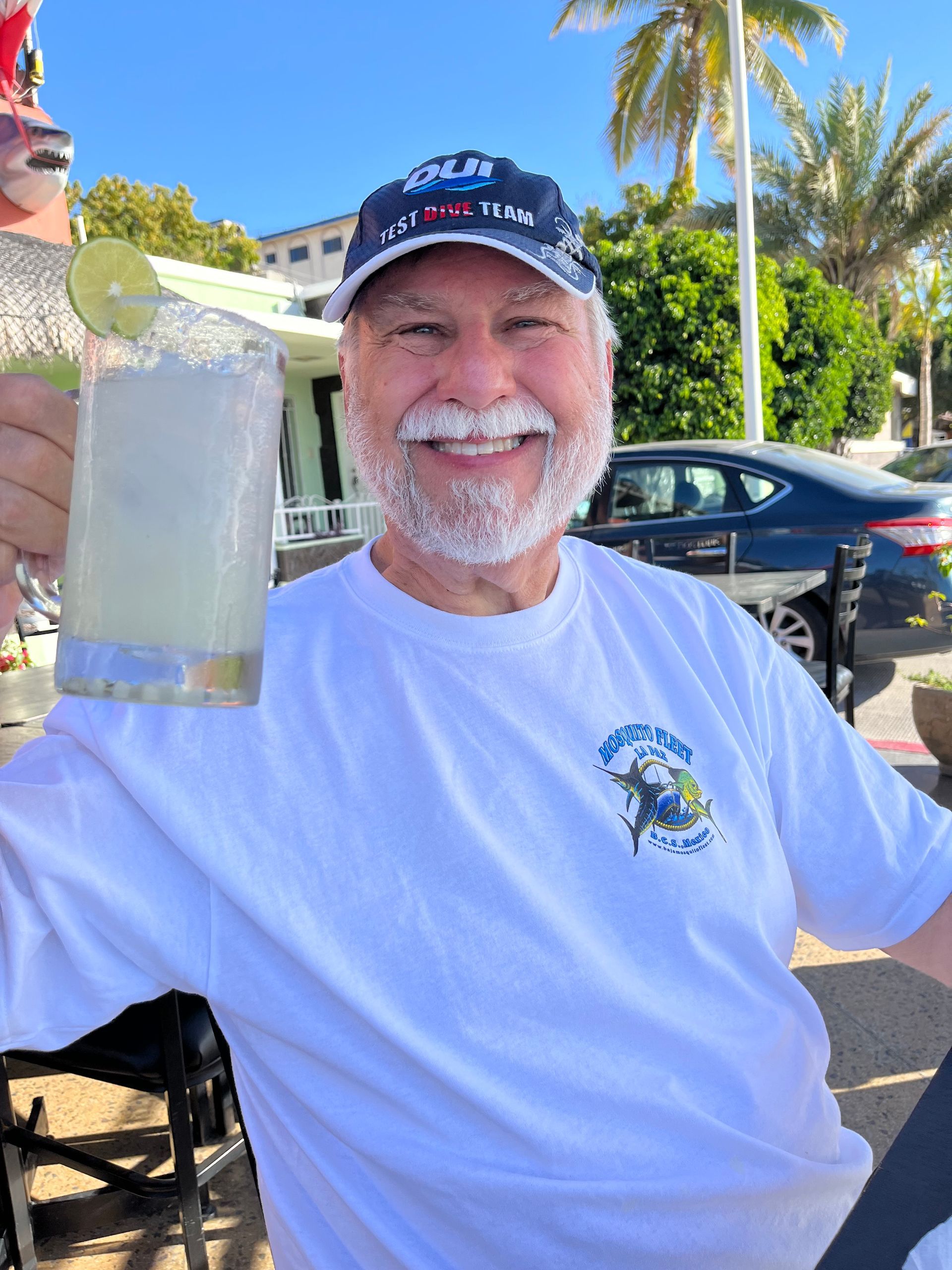 Sam Davis Board Member Man smiling in white t-shirt  and blue ballcap. holding up a drink