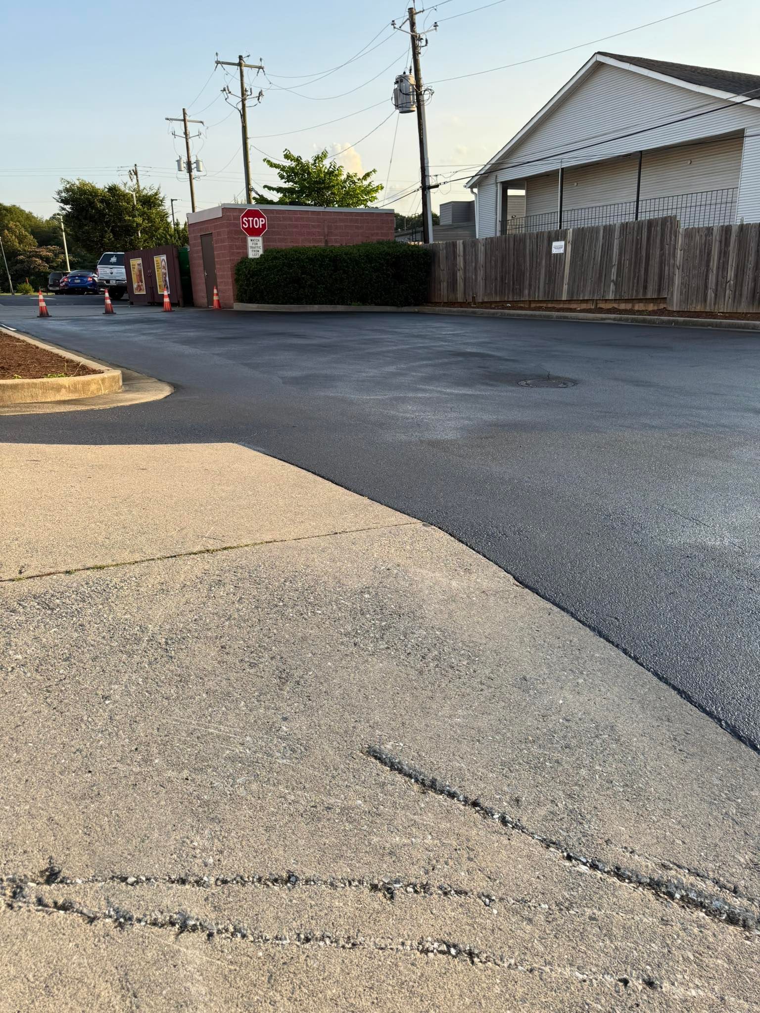 Asphalt road meets concrete sidewalk. Buildings and power lines in the background.