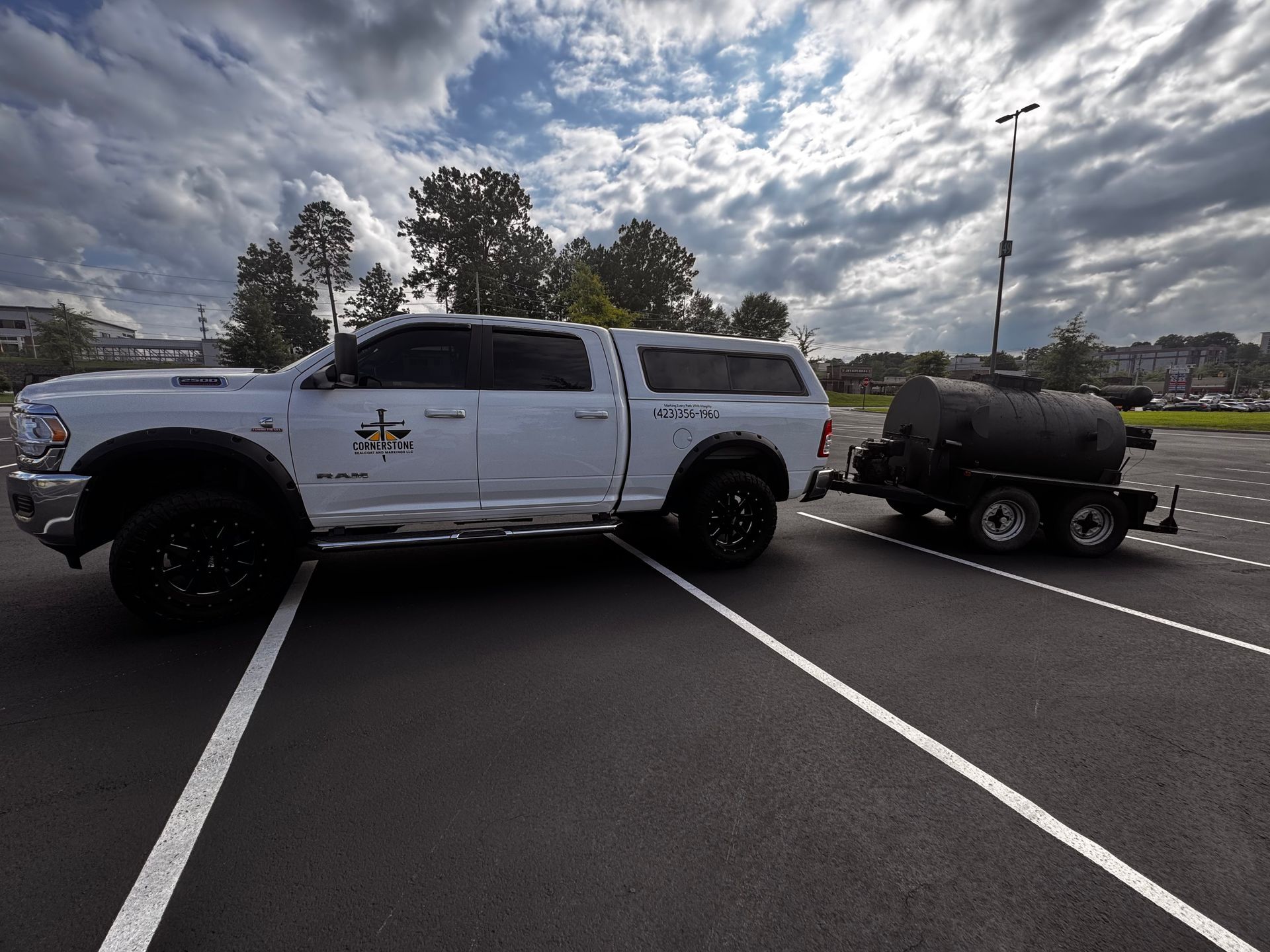 White pickup truck towing a trailer with a black smoker on asphalt parking lot under cloudy sky.