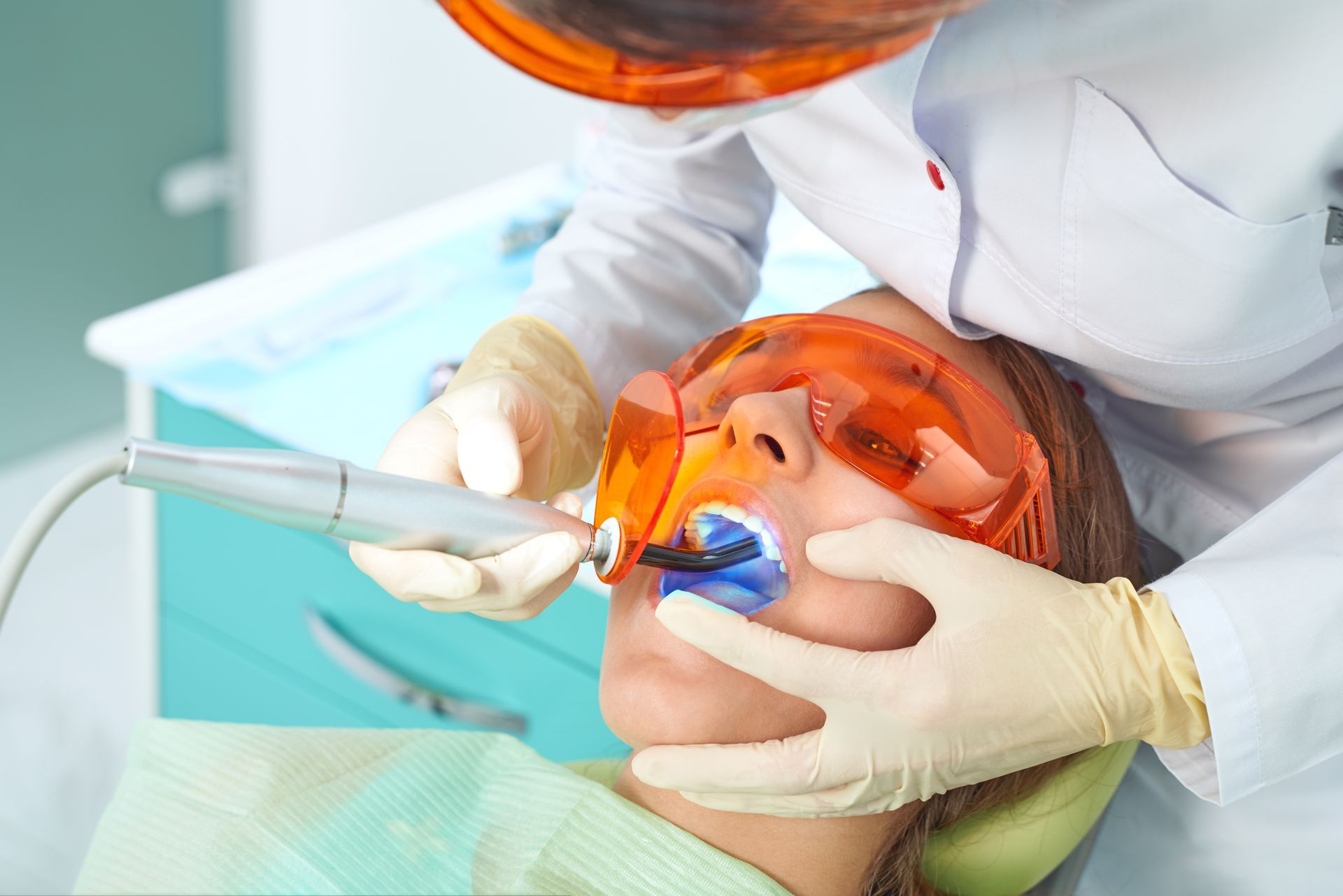 A dentist uses a blue light curing tool on a patient's teeth, both wearing orange-tinted protective glasses.