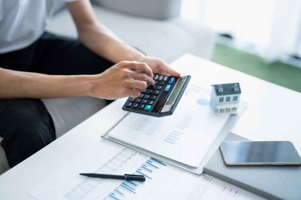 A person sitting at a desk uses a calculator next to a small model house, financial charts, a pen, and a smartphone.
