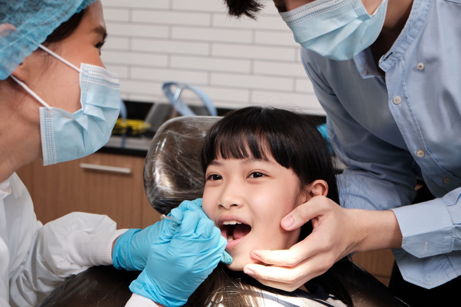 A child sits in a dental chair while a dentist performs a check-up, assisted by a person holding the child's cheek.