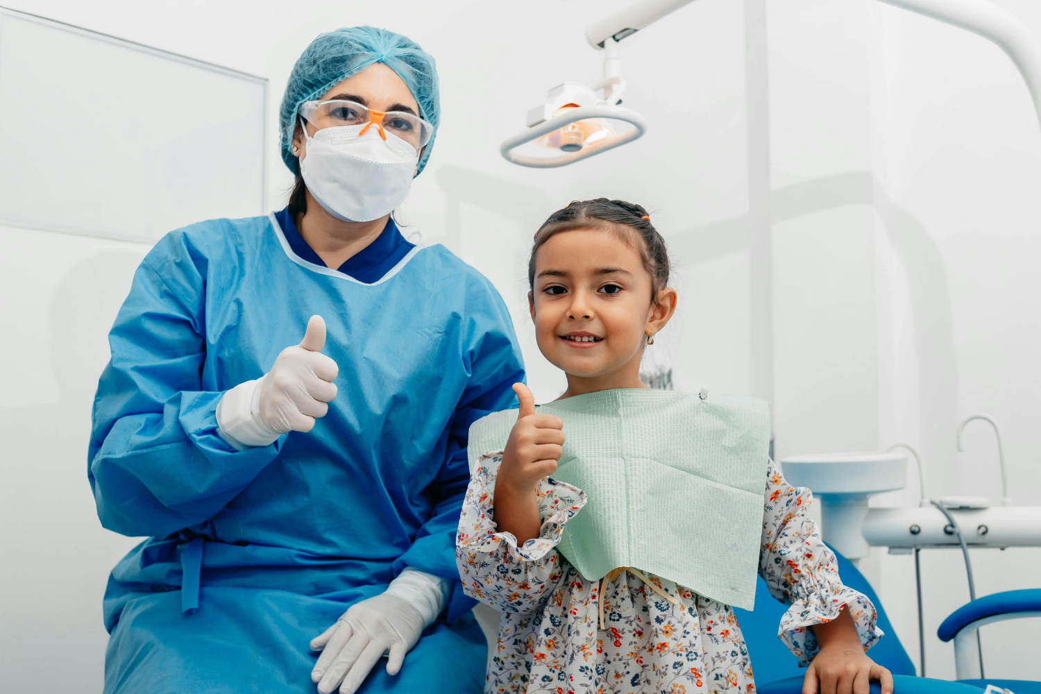 A dental professional and a child in a dental chair both give a thumbs-up in a clinical office.