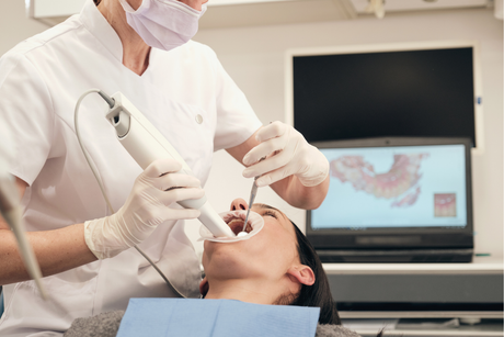 A dentist in a white coat and mask uses an intraoral scanner on a patient, with a 3D dental model displayed on a monitor.