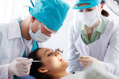 A dentist and assistant in protective gear perform a dental procedure on a patient in a bright clinical setting.