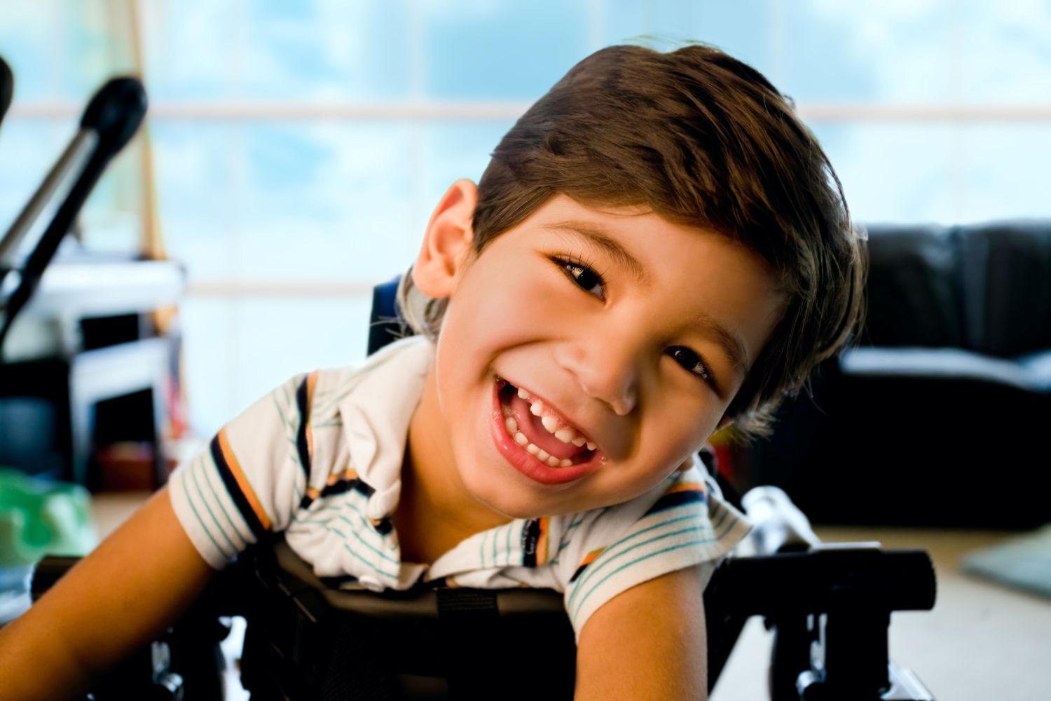 A smiling child with dark hair sits in a wheelchair, looking toward the camera in an indoor setting.