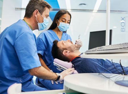 A dentist and an assistant wearing blue scrubs and masks examine a patient lying in a dental chair in a bright clinic.