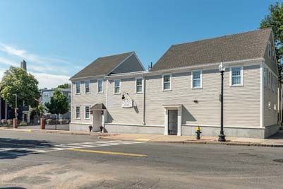 Two-story light gray building with multiple windows on a street corner under a blue sky.