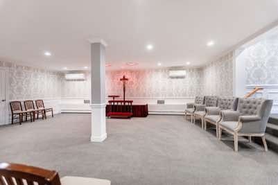Funeral chapel with a cross, chairs, and light gray carpet. White walls with floral wallpaper and recessed lighting.
