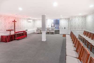 Interior of a funeral home with rows of chairs, a pulpit with a cross, and a doorway.
