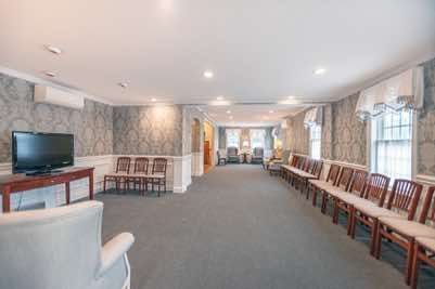 Funeral home waiting area with rows of chairs, TV, floral wallpaper, and large windows.