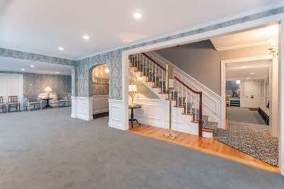 Spacious interior with staircase, grey carpet, wallpaper. Entryway to room on right and seating area on left.