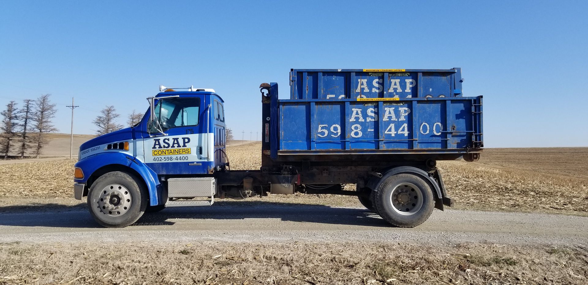 A blue ASAP containers dump truck is parked on the side of the road in a field.